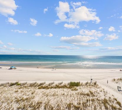 Pristine white sand beach stretches along turquoise waters under a brilliant blue sky dotted with fluffy white clouds.