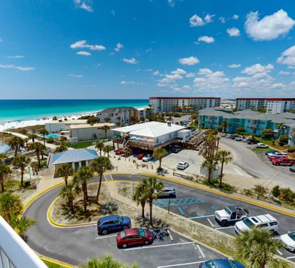 Beachfront property with stunning turquoise waters, white sand beach, and nearby hotels and shops. Palm-lined parking area marks the entrance, close to emerald Gulf shores.