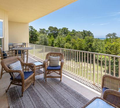 Expansive balcony overlooking lush treetops and distant sea views, featuring wicker seating with blue cushions and a dining table—perfect for morning coffee or sunset relaxation.