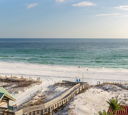 Pristine white sand beach stretches along emerald Gulf waters, with wooden boardwalk providing easy beach access beneath clear blue skies.