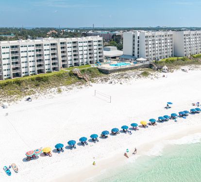 Aerial view of beachfront buildings with white sand beach, turquoise waters, and swimming pool visible in the resort complex.
