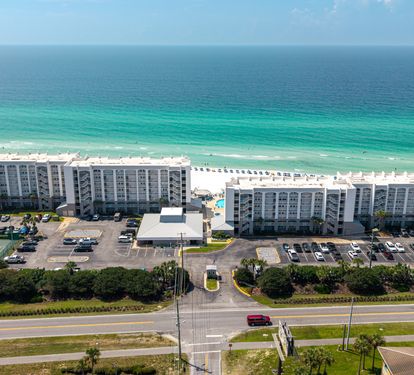 Aerial view of beachfront resort buildings with pristine white sand and emerald waters stretching to the horizon, surrounded by parking areas and coastal landscape.