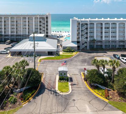 An elevated view of the beachfront property entrance, with the beach and Gulf visible just beyond the buildings and parking areas.