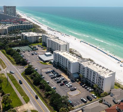 Aerial view of a beachfront property positioned along pristine white sand and turquoise waters, surrounded by coastal neighborhoods and convenient parking.