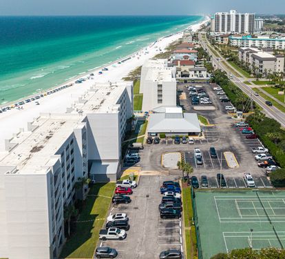 Beachfront property complex situated along pristine white sand shoreline with turquoise Gulf waters, featuring tennis courts, parking facilities, and convenient highway access to area attractions.