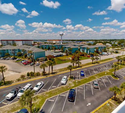 An aerial view of the vacation rental complex set among palm-lined streets and parking areas, with neighboring buildings and a water tower visible in the surrounding neighborhood.