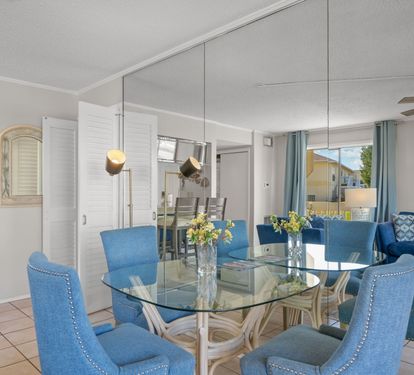 The dining area features a glass-top table with seating for four, coastal-inspired décor, and an open layout that flows seamlessly into the living space. A bright and inviting spot for meals, morning coffee, or gathering after a day at the beach.