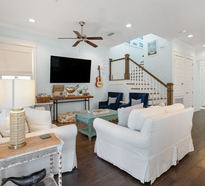 Sink into plush white sofas in your bright, coastal-styled living room where natural light flows through large windows and a ceiling fan keeps you comfortable.