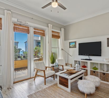 Light-filled living area with coastal flair, featuring comfy seating, a flat-screen TV, and glass doors that open to a palm-lined balcony—perfect for a laid-back 30A getaway.