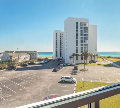 Beachfront view from the property window featuring nearby buildings, parking area, palm trees, and pristine turquoise ocean waters.