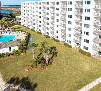 Aerial view of white oceanfront buildings with landscaped lawns, palm trees, and swimming pool, with water visible in the distance.