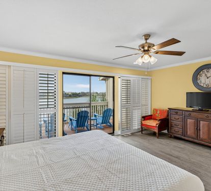 Wake up to waterfront views in your bright, comfortable bedroom with ceiling fan, TV, and private balcony access featuring blue Adirondack chairs.