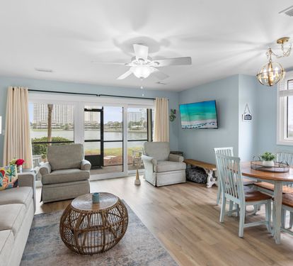 Cozy lakefront living room with armchairs, a rattan coffee table, flat-screen TV, and dining set for four. Sliding glass doors open to serene water views and a private patio.