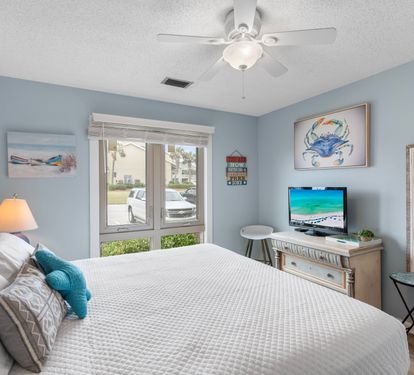 Bright coastal guest bedroom with a comfy king bed, beach-themed decor, flat-screen TV, and natural light pouring in through large windows—your perfect retreat after a day in the sun.
