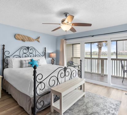 Spacious primary bedroom with a wrought iron king bed, coastal décor, and sliding doors that open to a private balcony with serene lake views—your perfect waterfront retreat.
