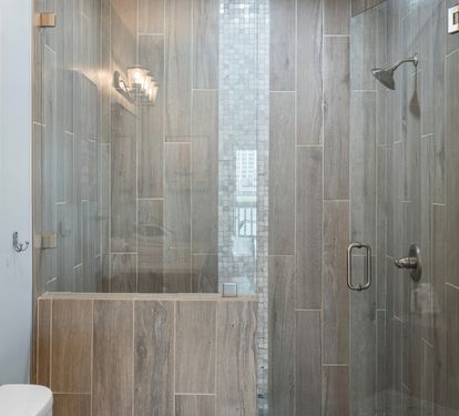 The en-suite bathroom features a luxurious walk-in shower with sleek wood-look tile, mosaic accent strip, and glass doors—blending spa-like comfort with modern coastal design.