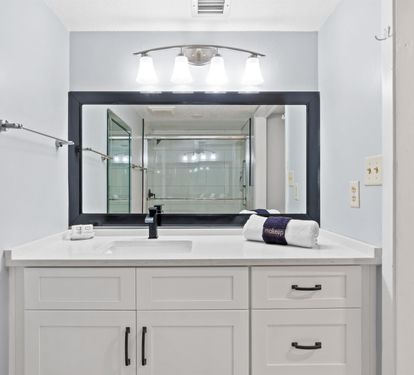 Your spacious bathroom features elegant white vanity with ample storage, modern lighting, and glass shower visible in the mirror reflection.