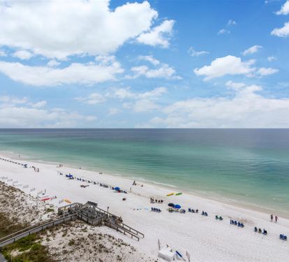 Pristine white sand beach stretches along emerald waters under a brilliant blue sky dotted with fluffy white clouds.