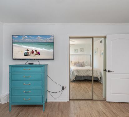 Your bright living space features a vibrant teal dresser and large TV, with glimpses of your cozy bedroom through mirrored doors.