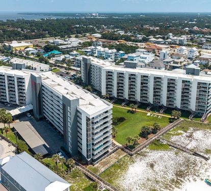 Aerial perspective of the beachfront property complex and surrounding coastal community, showcasing the building's prime location near white sandy shores and nearby waterfront areas.
