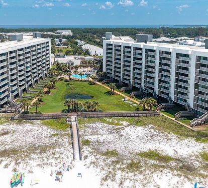 This aerial view captures two modern beachfront buildings flanking a landscaped courtyard with a sparkling pool and lush palm trees, all set along a pristine white sandy beach.