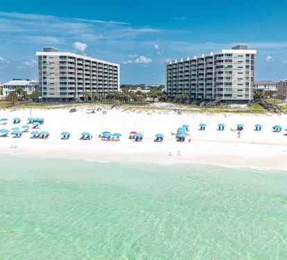 A stunning beachfront property with direct access to pristine white sand and crystal-clear turquoise waters, where colorful umbrellas dot the shore under endless blue skies.