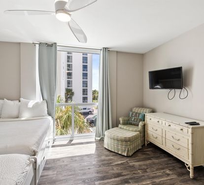Your coastal bedroom features plush bedding, ceiling fan comfort, and bright city views through floor-to-ceiling windows—perfect for restful nights and refreshing mornings.
