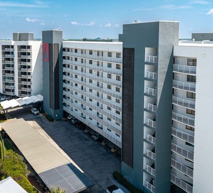 An aerial perspective showcasing the multi-story building exterior, complete with covered parking and landscaped palm trees. The property is set within a developed area under bright, sunny skies.